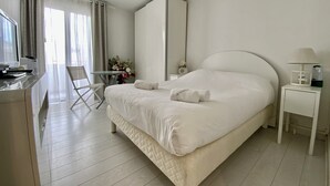 A hotel room with a large bed, a desk, a chair, a small table with flowers, and a nightstand with a lamp.