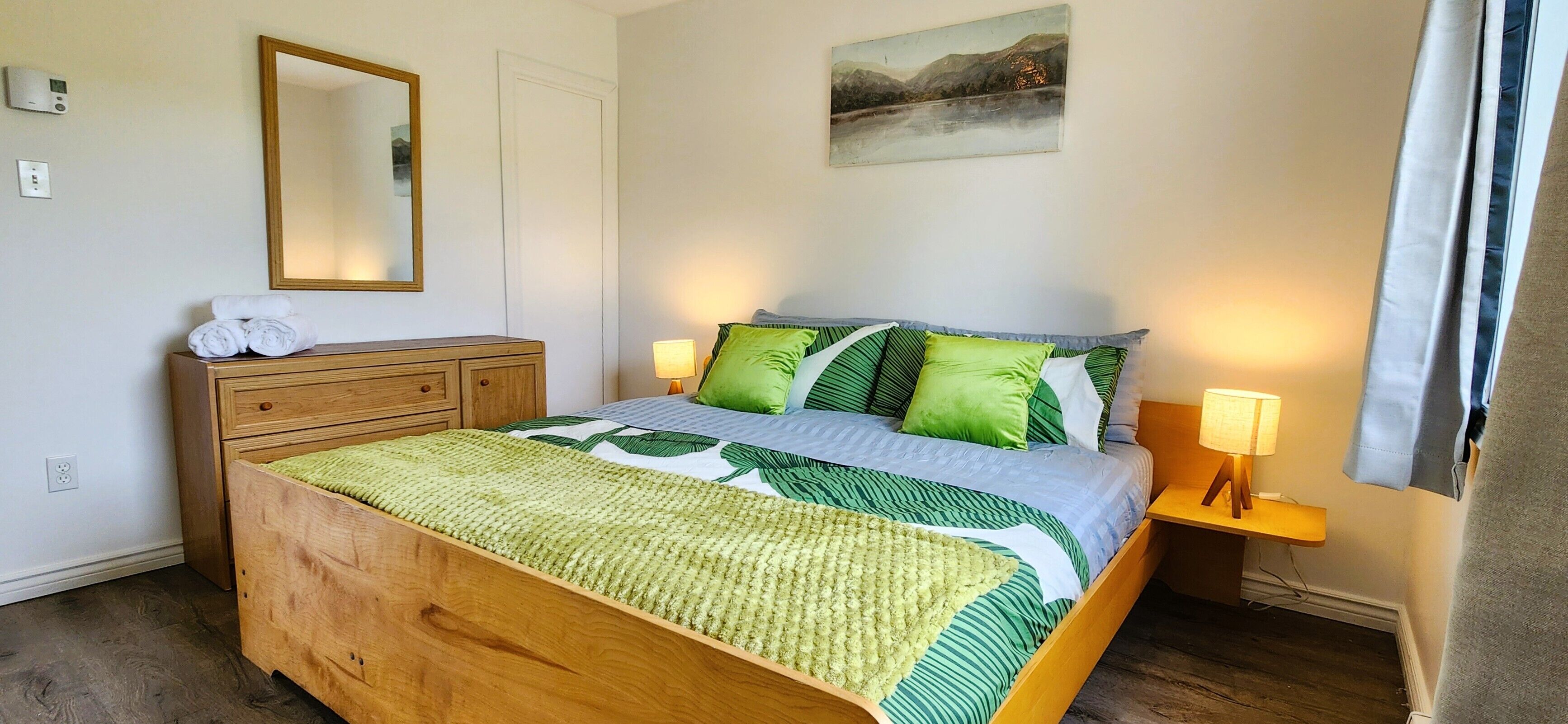 Une chambre à coucher avec un lit en bois, une literie verte et blanche, une lampe de chevet, un miroir et un tableau accroché au mur.