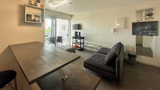 A modern living room with a sofa, a dining table, and a TV. There is a balcony with a chair and a plant.