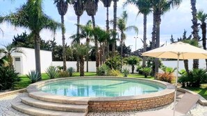 Una piscina con borde de ladrillo, rodeada por un patio de mosaicos, palmeras y un muro blanco al fondo.