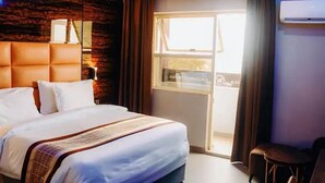 A hotel room with a large bed, a wooden headboard, a window with curtains, and an air conditioning unit. - San Antonio Blu Hotels (Awka)