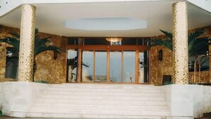 A hotel entrance with a set of stairs leading up to a glass door, flanked by two columns with textured stone patterns. - San Antonio Blu Hotels (Awka)