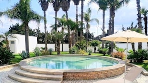 A swimming pool with a brick border, surrounded by a mosaic tile patio, palm trees, and a white wall in the background.