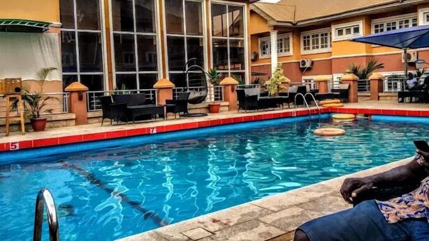 A swimming pool with a tiled edge, surrounded by outdoor seating and a building with large windows. - Hallydays Hotel and Resort (Makurdi)