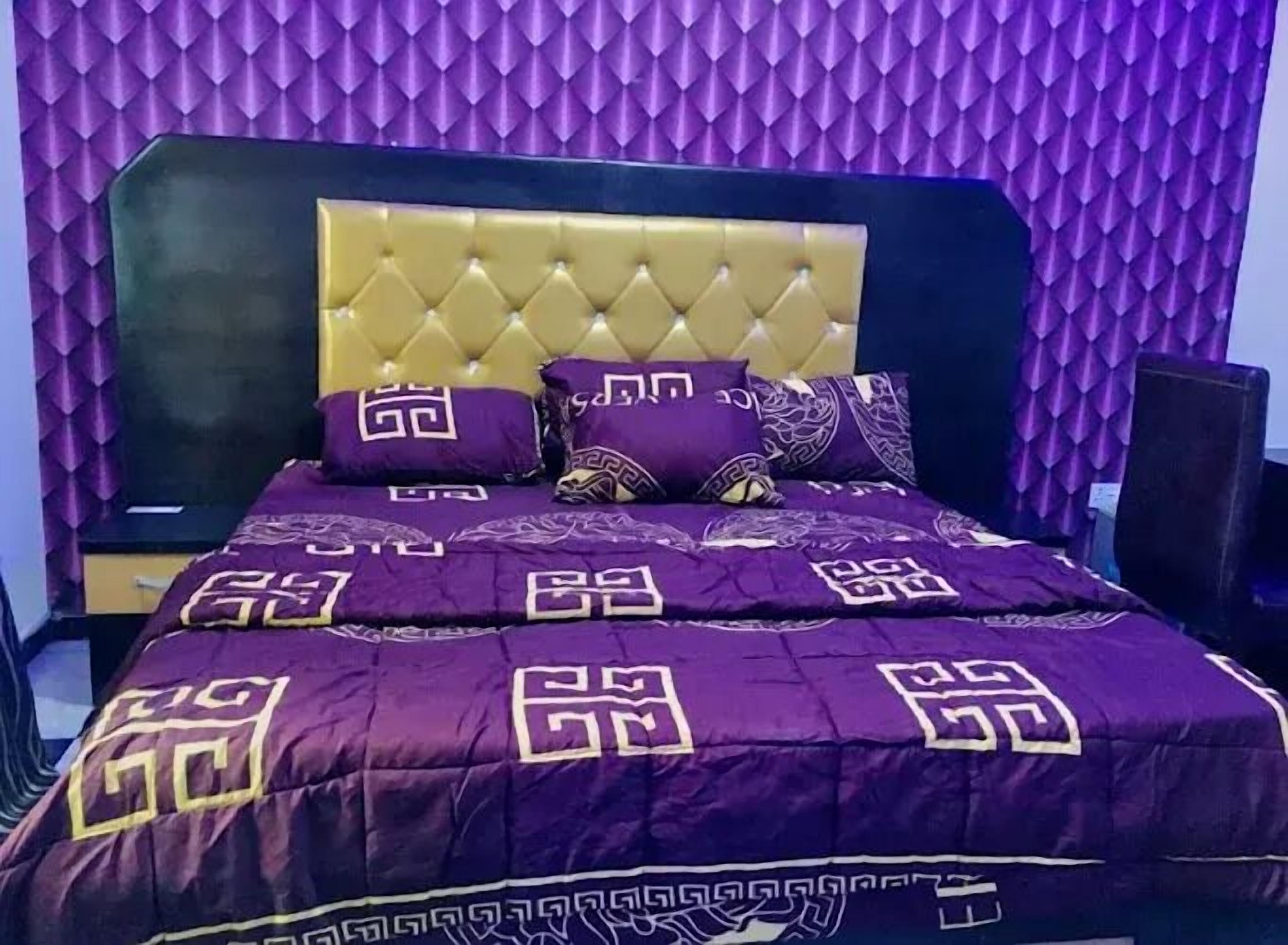 Un dormitorio con cabecera capitonada, ropa de cama morada con diseños geométricos y una pared texturizada color púrpura.