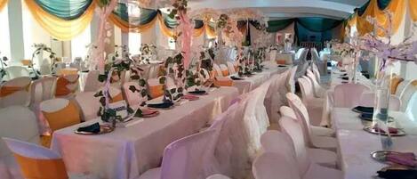 Un salón de banquetes con mesas largas, sillas blancas y centros de mesa florales.