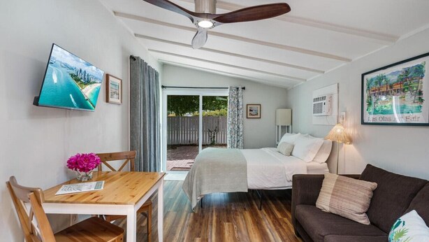 Habitación de hotel con cama, escritorio, sillón, sofá, ventilador de techo y televisor en la pared.
