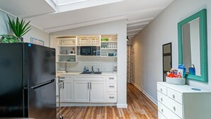 Cocina compacta con armarios blancos, un refrigerador negro y un microondas. Tiene piso de madera y una pequeña zona de comedor con una silla.
