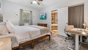 Un dormitorio con cama, un escritorio con una computadora portátil, un ventilador de techo y un baño con ducha y estantes.