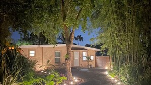 Una casa con entrada pavimentada, rodeada de árboles y plantas, e iluminada por luces en el suelo.