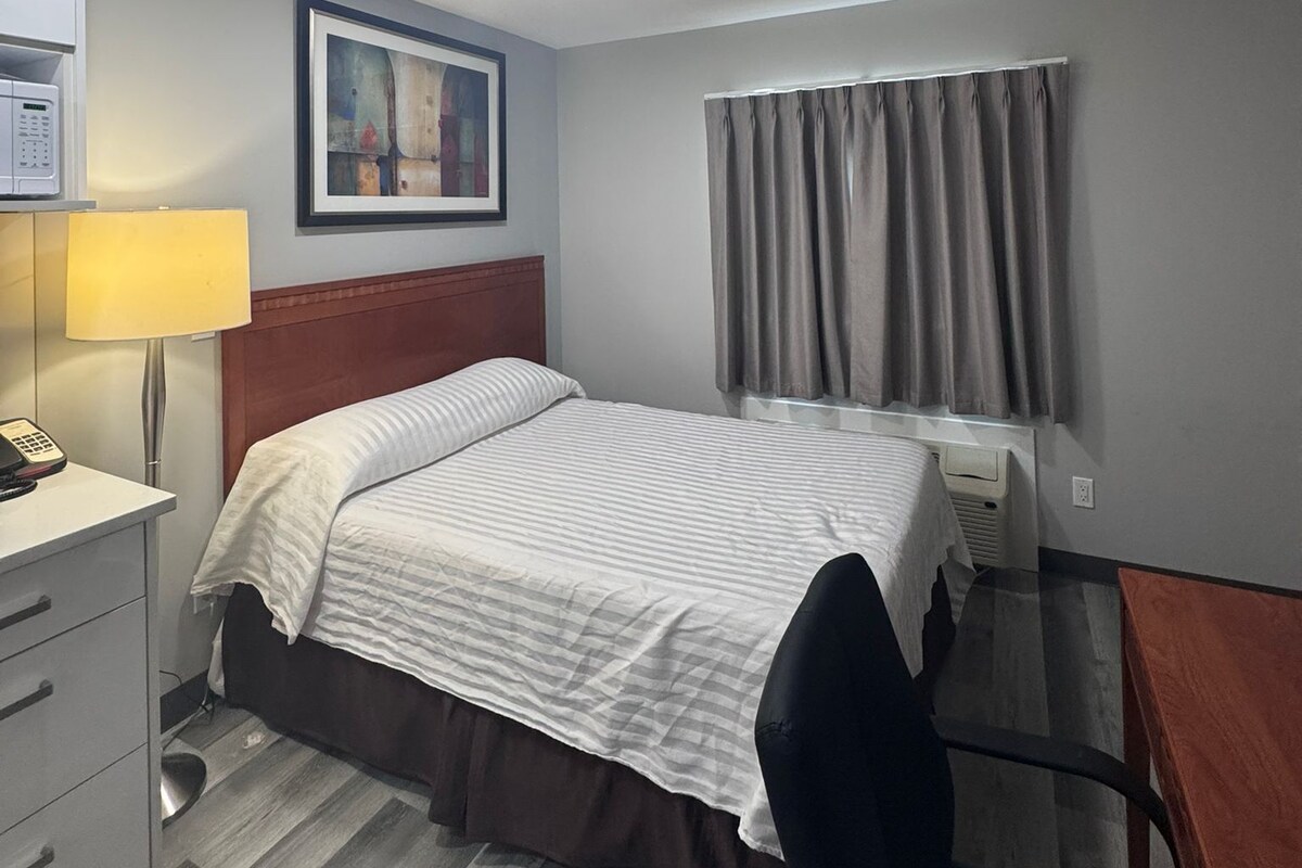A hotel room with a bed, a desk, a lamp, a painting, and a window with curtains.
