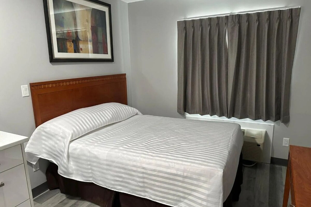 A neatly made bed with a wooden headboard, a gray curtain covering a window, and a framed abstract painting on the wall.
