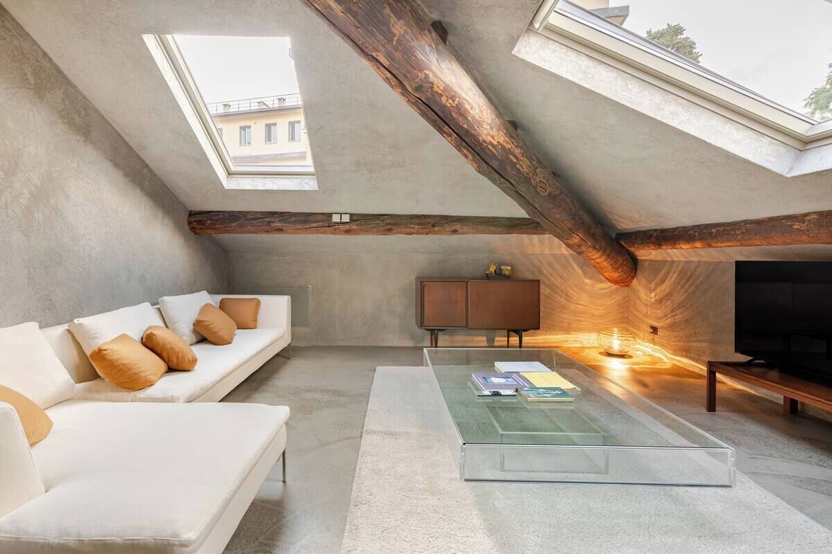 An attic living room with a high ceiling, exposed wooden beams, a white sofa, a glass coffee table, and a television.