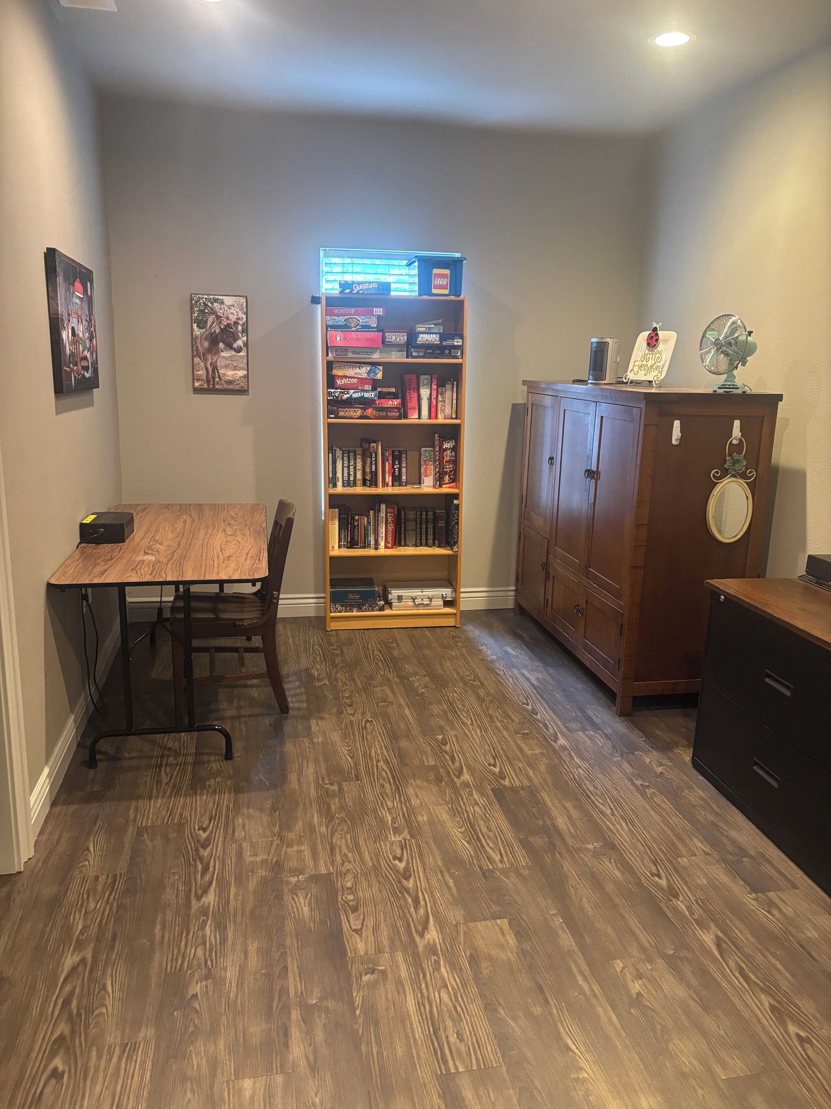 A room with a wooden table, chairs, a bookshelf, a cabinet, and a fan.