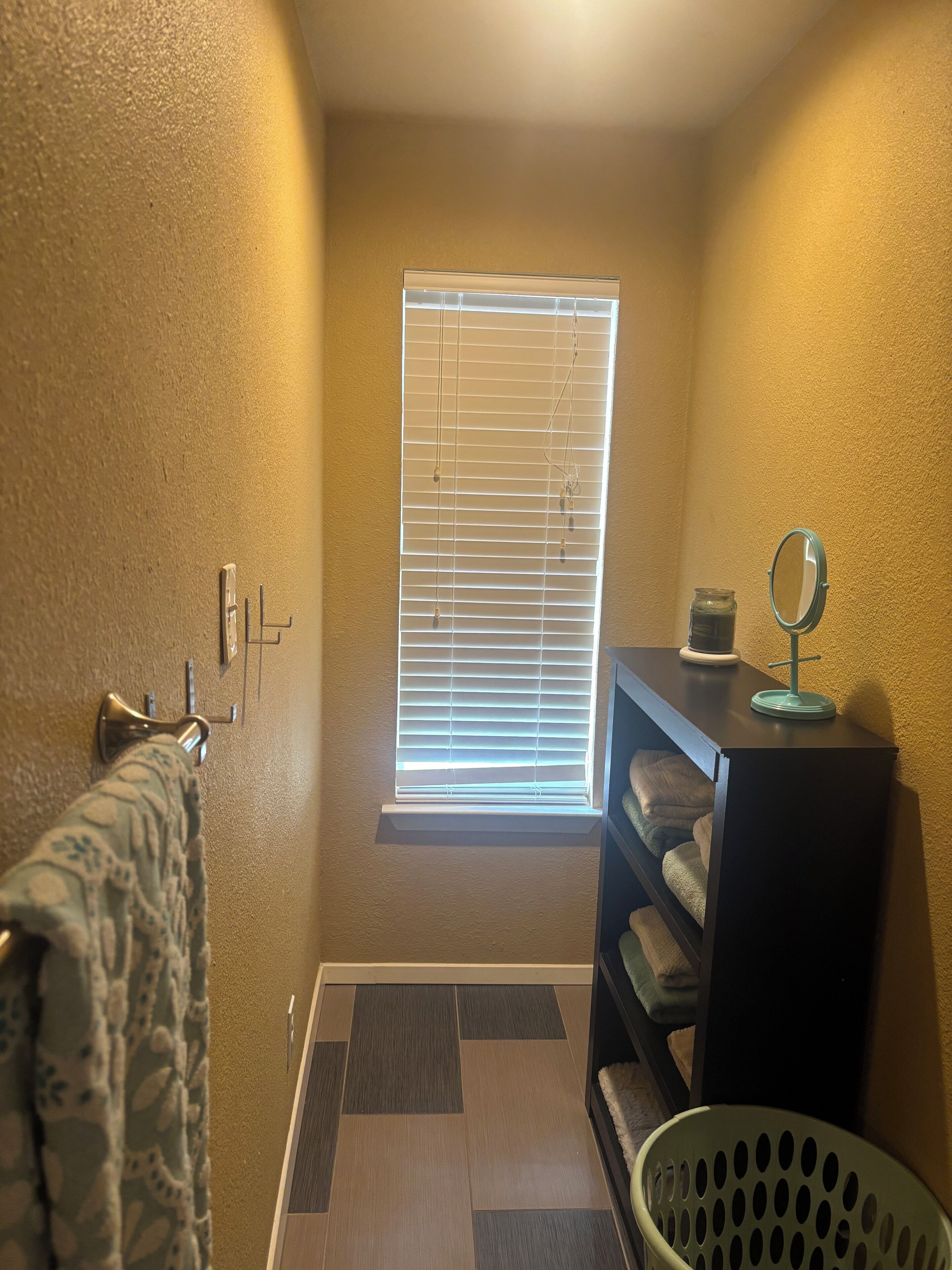 Un baño pequeño y estrecho con una ventana, un perchero, un estante con un espejo y un cesto de ropa.