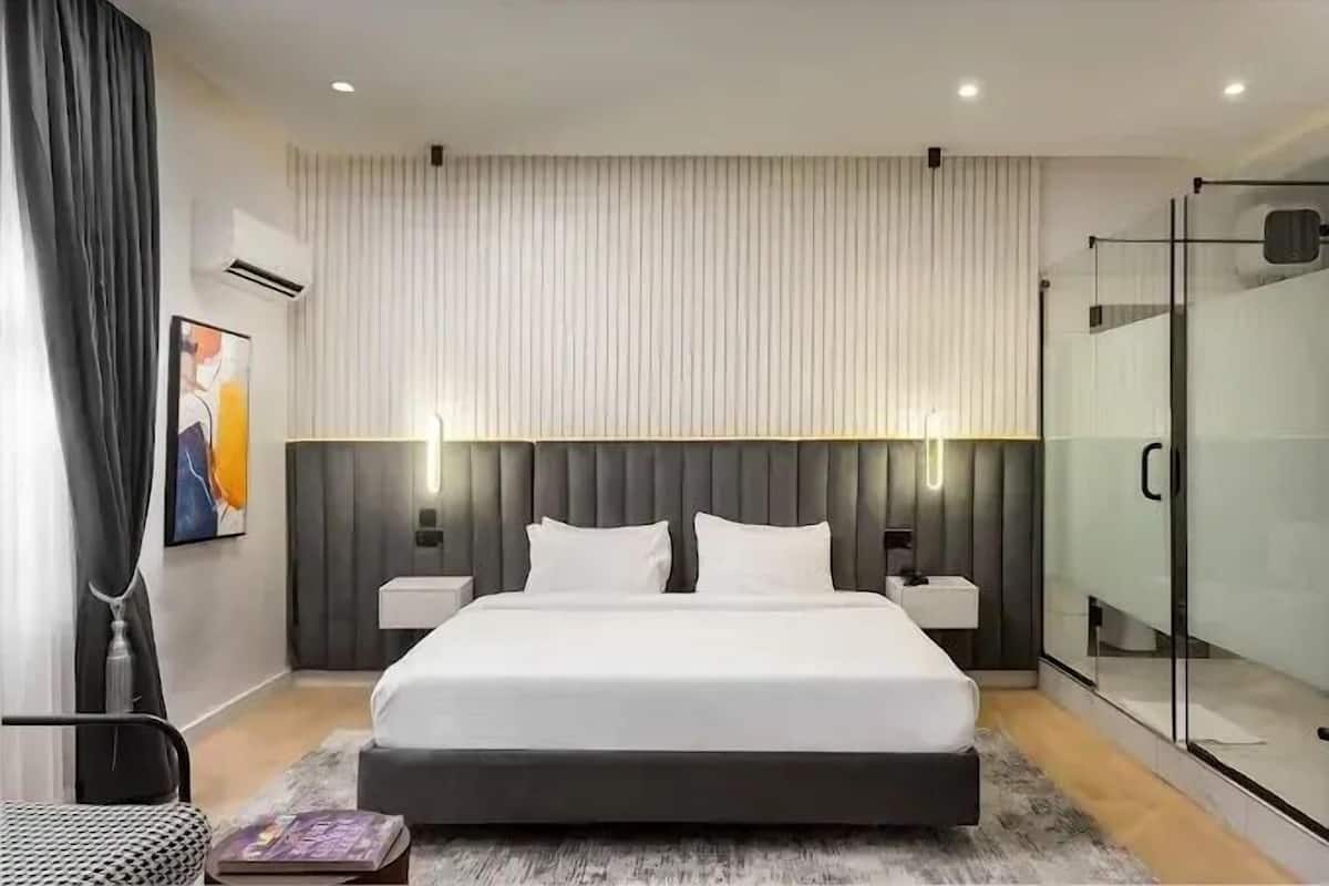 A modern hotel room with a large bed, bedside tables, a glass shower enclosure, and a view of the city.