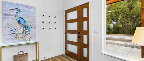 Un pasillo con suelo de madera, una puerta con paneles de vidrio, una cesta fijada a la pared y un cuadro enmarcado de un pájaro.
