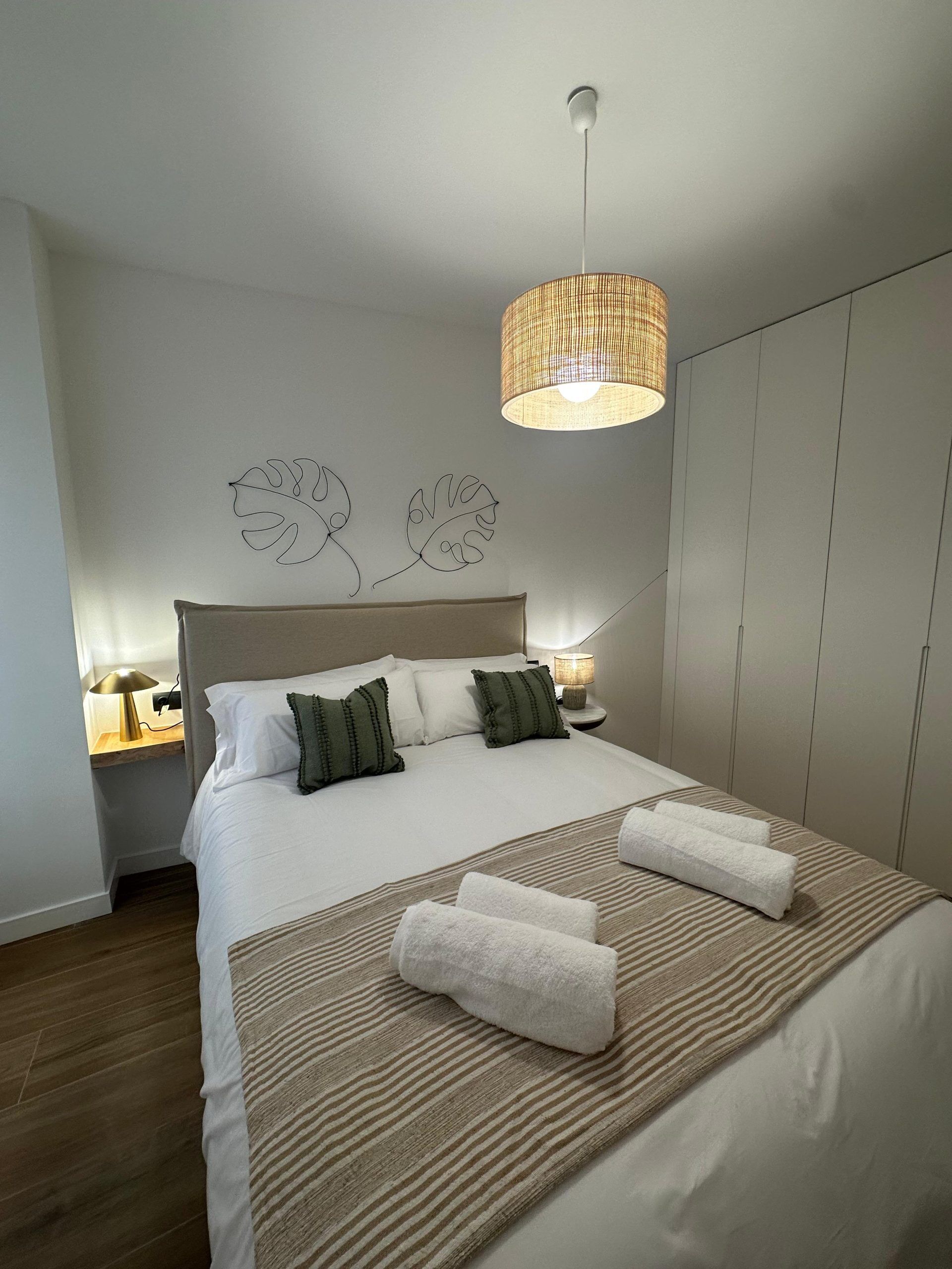 Una cama bien hecha con sábanas blancas y almohadas verdes, una mesita de noche con una lámpara y un candelabro colgante sobre la cama.