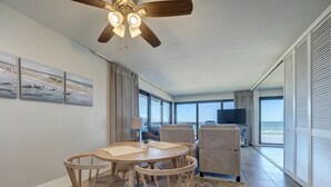 A modern living room with a round wooden table, four chairs, a ceiling fan, and a large window offering a view of the ocean. - Corner Beachfront Condo: Stunning 270° Views. Wraparound Balcony. Heated Pool (Corpus Christi)