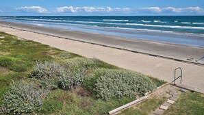 A coastal road with a beach in the background, a sidewalk, and some vegetation. - Corner Beachfront Condo: Stunning 270° Views. Wraparound Balcony. Heated Pool (Corpus Christi)