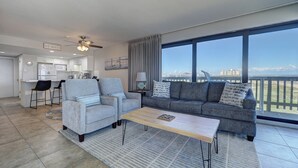 A modern living room with a sofa, two armchairs, a coffee table, and a view of the city through large windows. - Corner Beachfront Condo: Stunning 270° Views. Wraparound Balcony. Heated Pool (Corpus Christi)