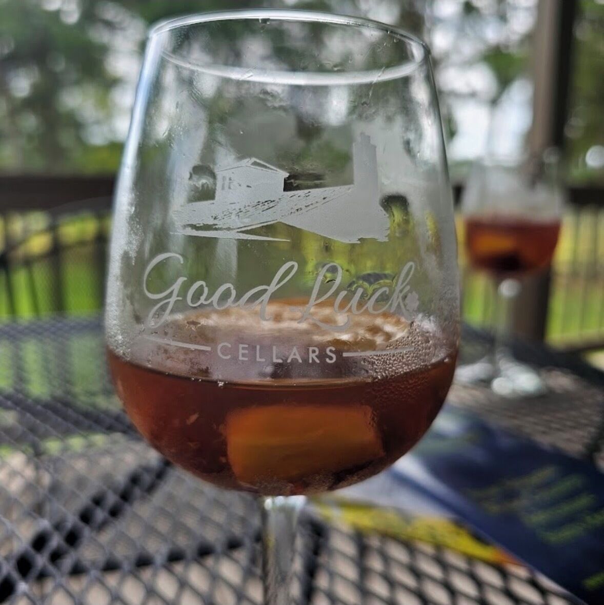 A glass with a logo and the text "Good Luck Cellars" on a metal table outdoors.