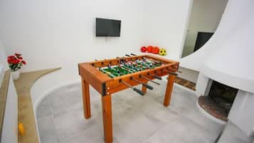 Une table de baby-foot dans une pièce dont le sol est carrelé, avec un téléviseur à écran plat accroché au mur et une cheminée.