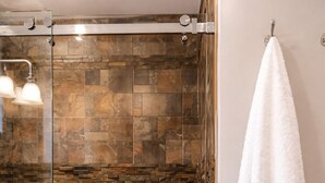 Ducha con puerta de vidrio, baldosas de piedra, una toalla blanca en un perchero y un barril de madera con velas.