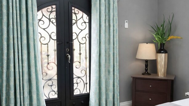 Un dormitorio con cama, mesita de noche, lámpara, planta en maceta y puerta con cortinas decoradas.