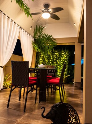 Un área de comedor al aire libre con sillas de mimbre y cojines rojos, un ventilador de techo y una pared decorada con luces y follaje.