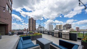 Terraza en la azotea con mobiliario exterior, una parrilla y luces colgantes, con el horizonte de la ciudad de fondo.