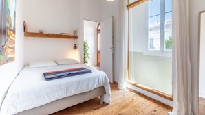 Un dormitorio con un ventanal grande, suelo de madera, una cama con ropa blanca, una lámpara colgante y un estante pequeño con libros y una planta.