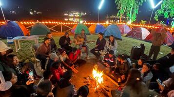Eine Gruppe von Menschen versammelt sich an einem Lagerfeuer in einer Outdoor-Umgebung, mit Zelten und Bäumen.