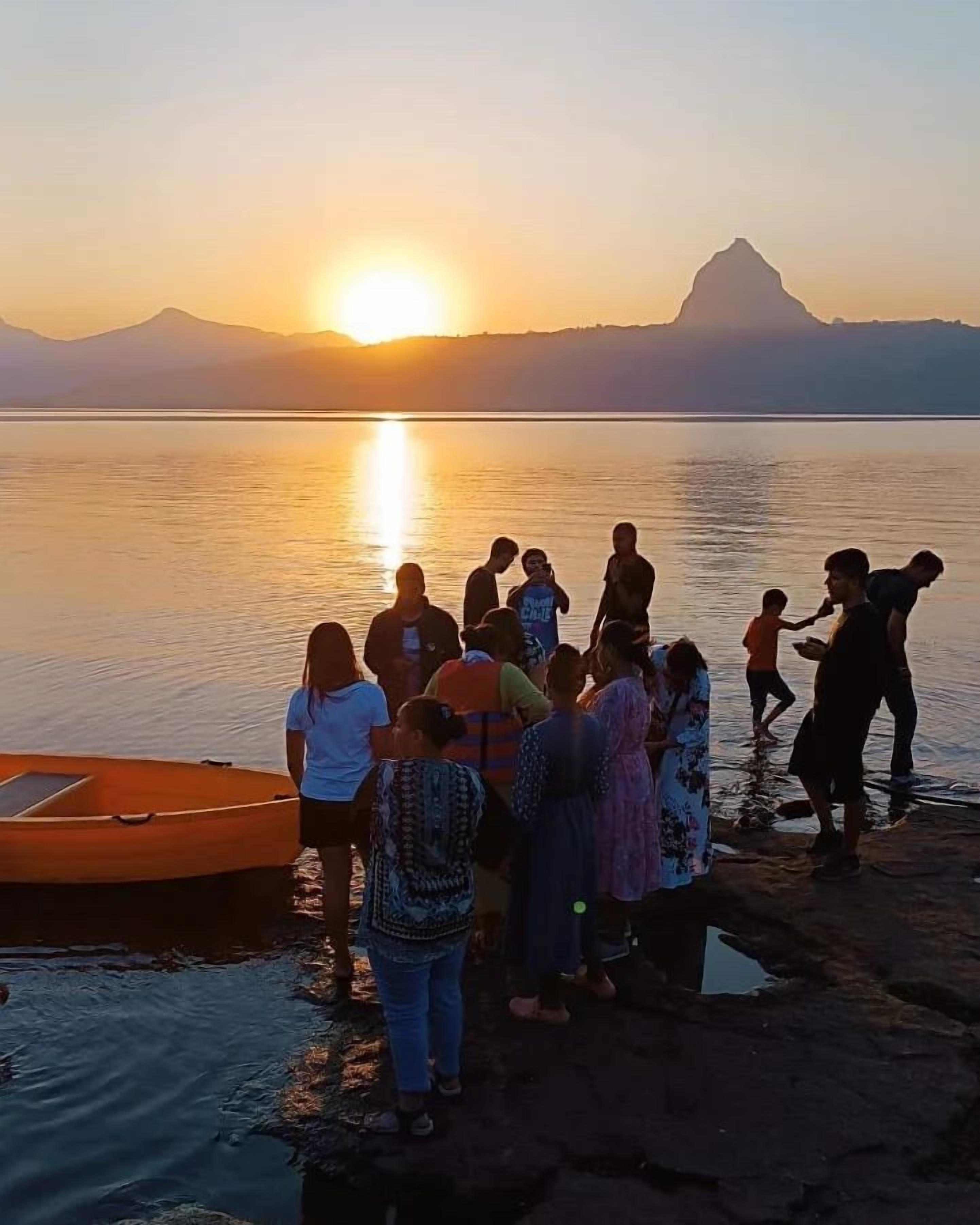 A group of people standing by a body of water at sunset with mountains in the background.