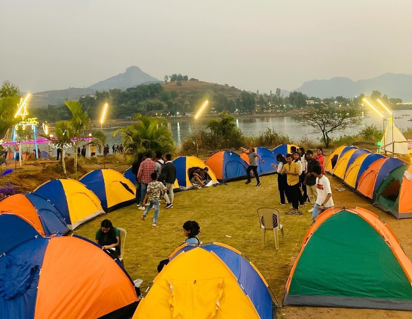 Un camping con varias tiendas de campaña dispuestas en filas, un lago al fondo y una colina con árboles.