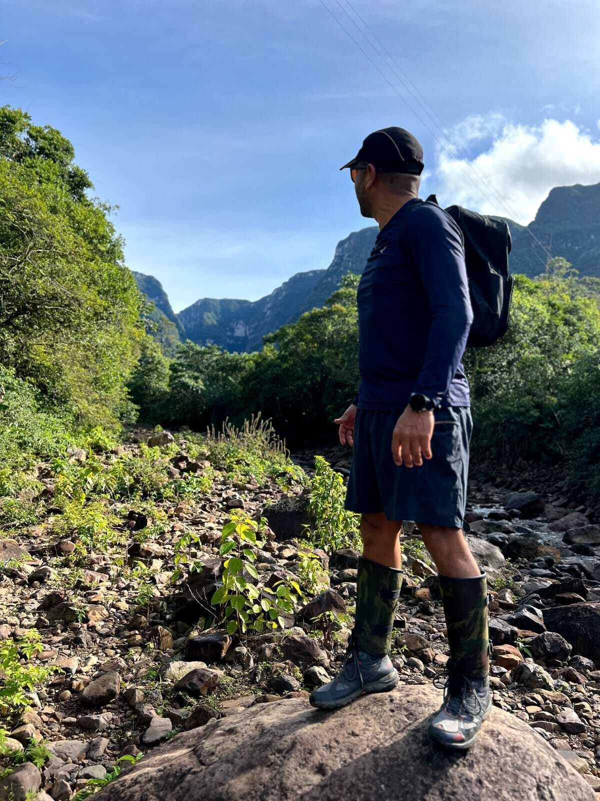 Una persona camina por un sendero rocoso con una mochila, rodeada de exuberante vegetación y montañas al fondo.