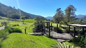 A rustic wooden structure with a thatched roof, surrounded by lush green grass and trees, set against a backdrop of mountains.