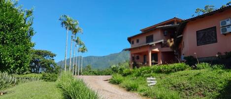 A rural house with a dirt path leading to it, surrounded by greenery and palm trees.