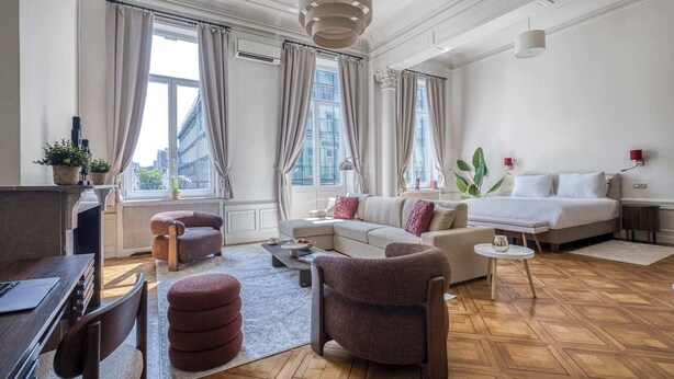A modern hotel room with a sofa, armchair, and a bed. There is a desk with a computer and a lamp. The room has large windows and a view of the city. - Deluxe Loft Mas C-12 (Brussels)