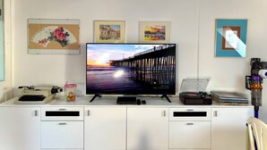Une pièce avec un téléviseur à écran plat posé sur un meuble blanc, une machine à écrire ancienne, un bocal rempli de fruits et des œuvres d’art encadrées accrochées au mur.