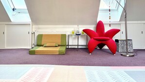 Une pièce avec un plafond haut, des fenêtres de toit, un canapé vert, un fauteuil rouge, un lampadaire et un tapis à motifs.