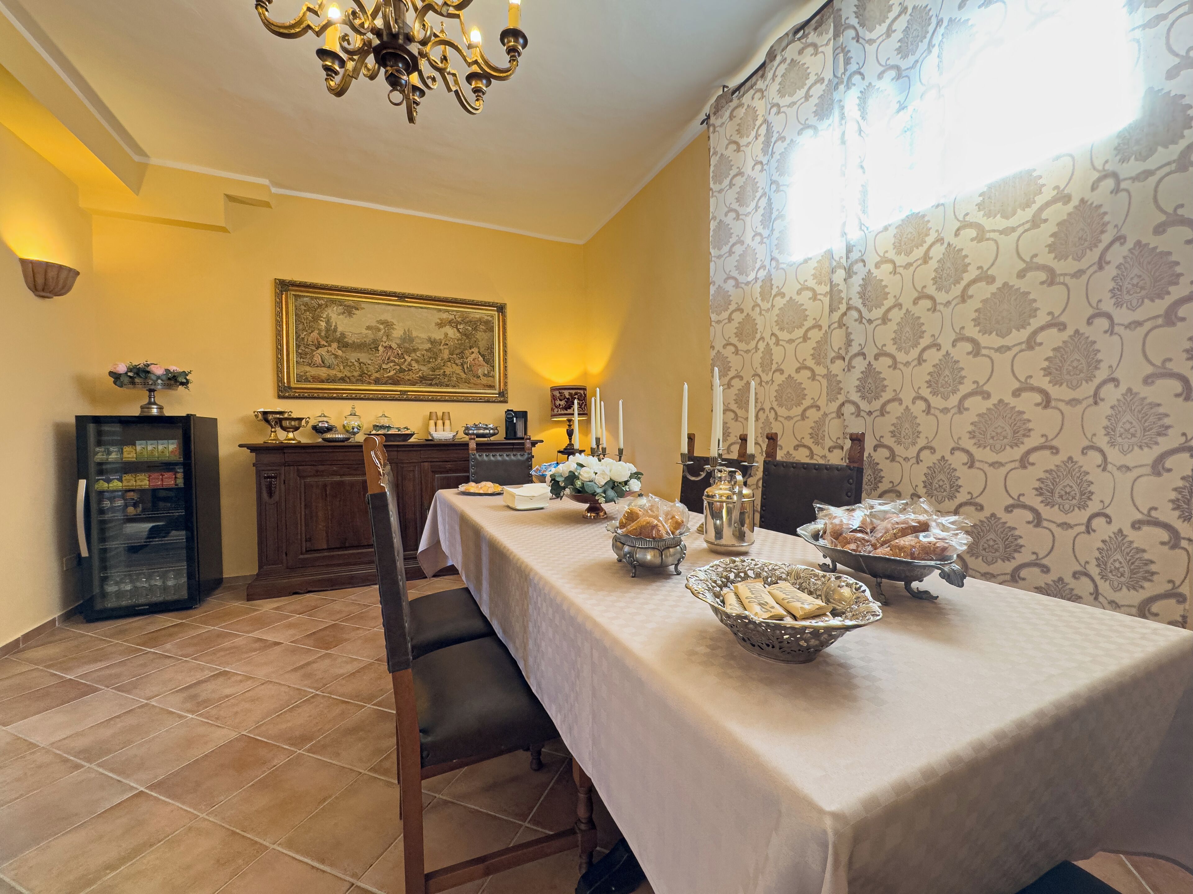 Un comedor con una mesa larga dispuesta para una comida, una araña, un aparador con objetos decorativos y un papel tapiz estampado.