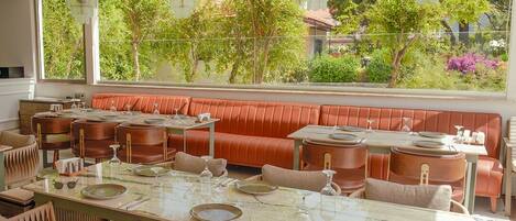 Un restaurante con una larga mesa dispuesta para comer, asientos con cojines y vistas a espacios verdes.