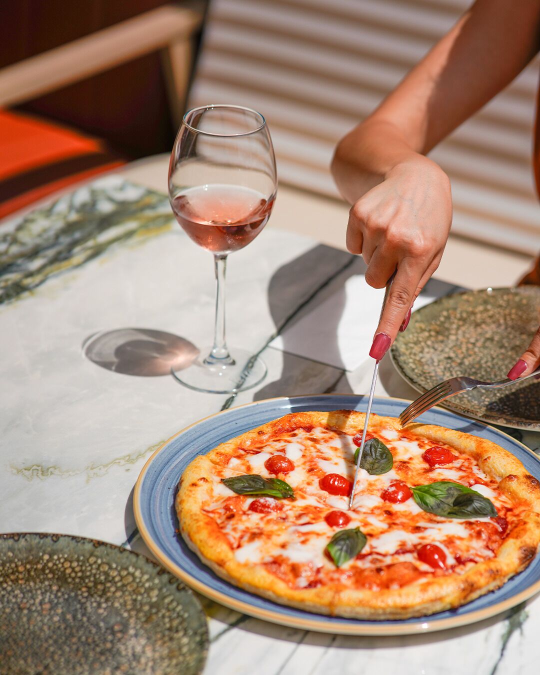 Une pizza servie sur une assiette, accompagnée d’une fourchette et d’un couteau, un verre de vin rouge et une table au motif de marbre.