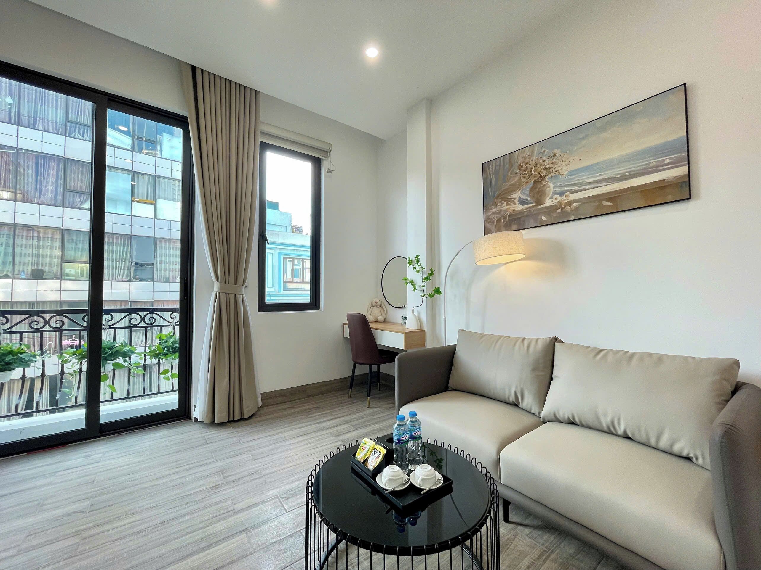 A hotel room with a sofa, a round table with a tray of items, a desk with a chair, a large window with curtains, and a painting on the wall.