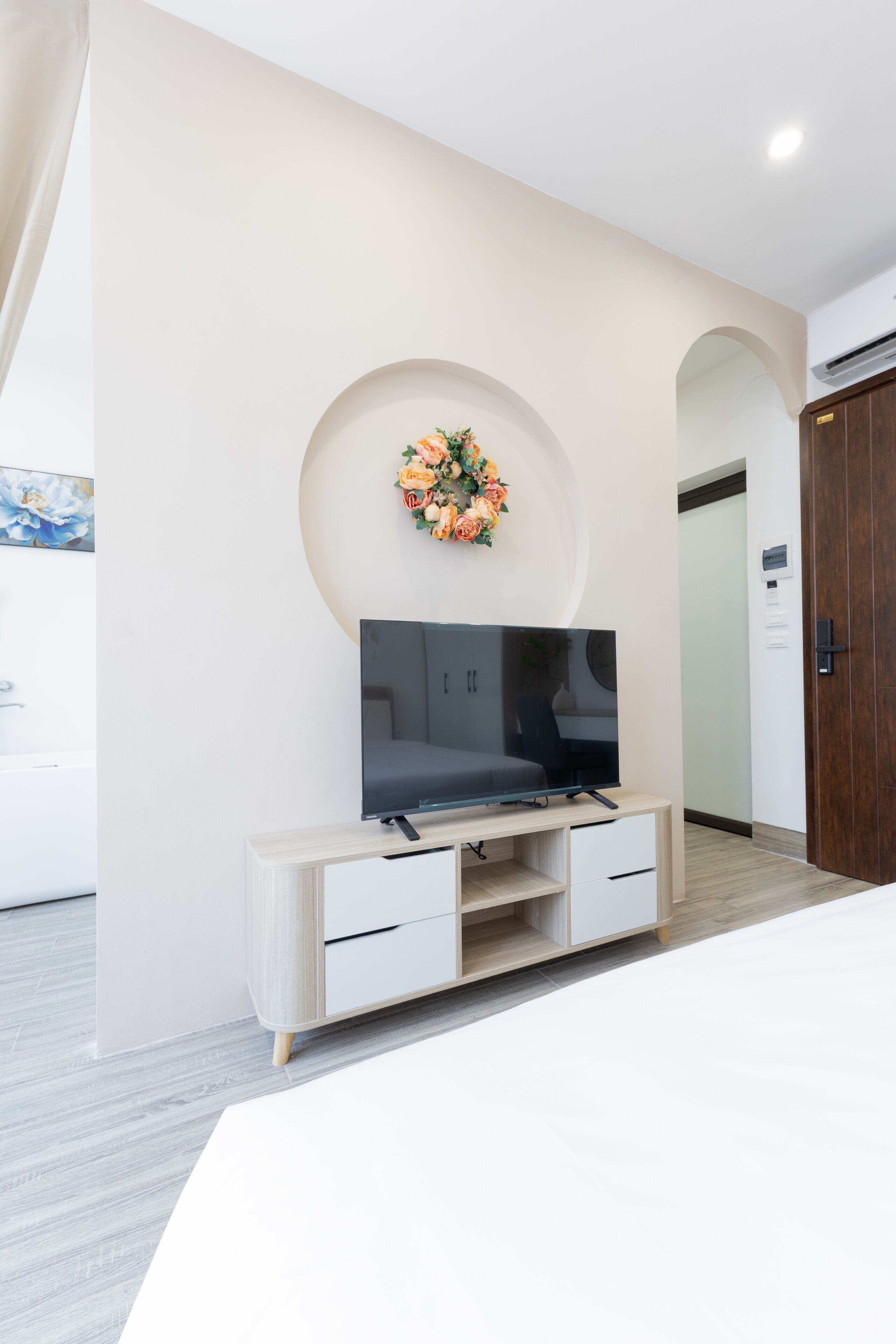 A modern hotel room with a flat-screen TV mounted on a wooden cabinet, a decorative floral arrangement on the wall, and a wooden door.