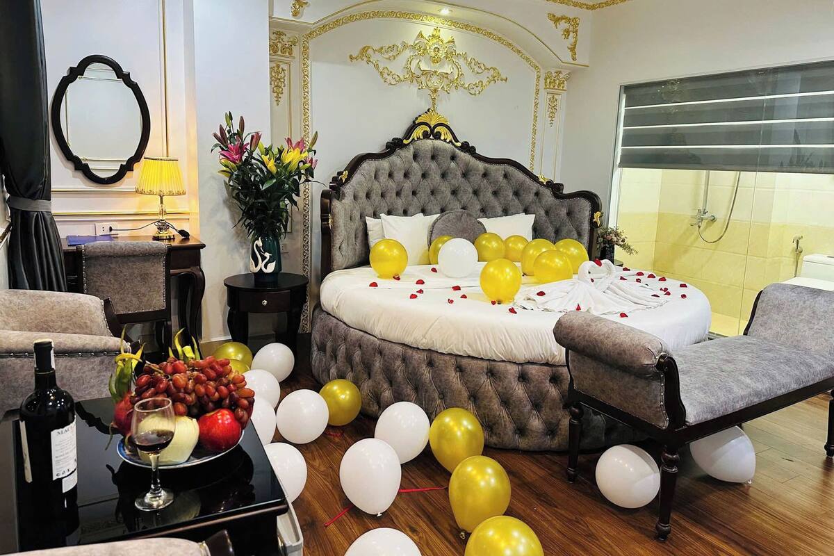 Ein luxuriöses Schlafzimmer mit einer gepolsterten Kopfteil, einem großen Bett mit weißen und gelben Ballons, einer Sitzecke mit einem Sessel, einem kleinen Tisch mit einer Weinflasche und Früchten sowie einem Spiegel an der Wand.