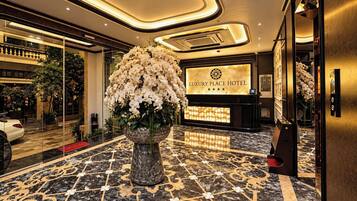 Zona de recepción de un hotel con suelo de mármol, un arreglo floral grande y un mostrador de recepción con un letrero que dice 'LUXURY PLACE HOTEL'.