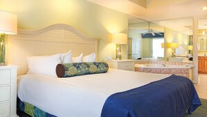 Habitación de hotel con una cama grande, una mesita de noche, un espejo, un baño con lavamanos y bañera, y una puerta que da a otra habitación.