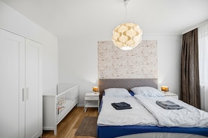 A modern bedroom with a white wardrobe, a crib, a bed with a blue bedspread, and a brick-patterned headboard.